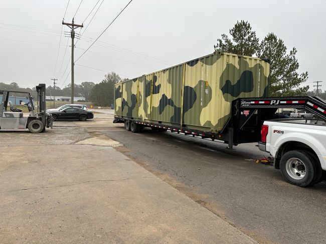 A shipping container custom painted with a a camo pattern loaded on a trailer.